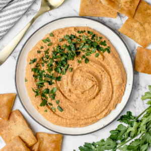 Butter Bean Hummus dip topped with fresh parsley and cracked black pepper. A spoon, extra parsley, and pita chips served on the side.