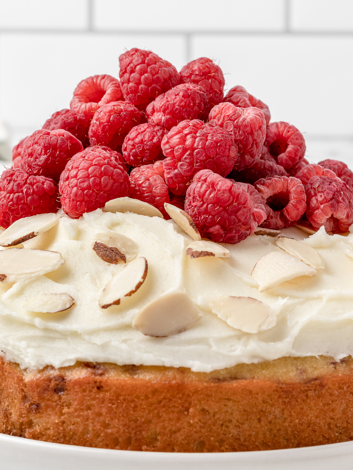 Close up of cake topped with almond frosting, almond slices, and a pile of fresh raspberries.
