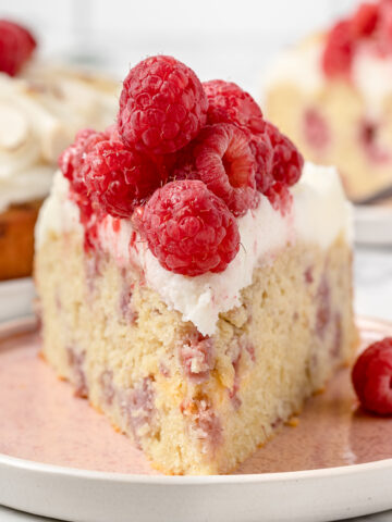 Close up of a slice of raspberry almond cake on plate to see tender crumb of the cake, creamy frosting, and plump raspberries on top.