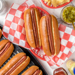 Smoked Hot Dogs in a basket ready to be topped with toppings and more hot dogs in buns on the side.