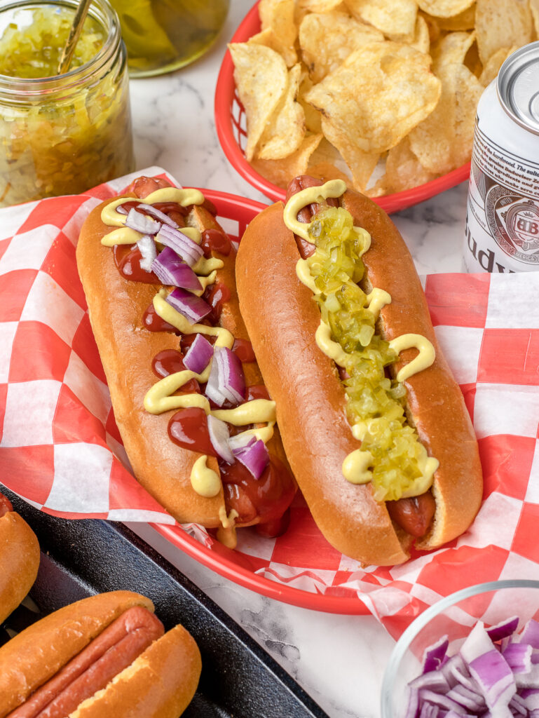Smoked hot dogs in brioche buns topped with toppings, mustard, ketchup, relish, and red onion.