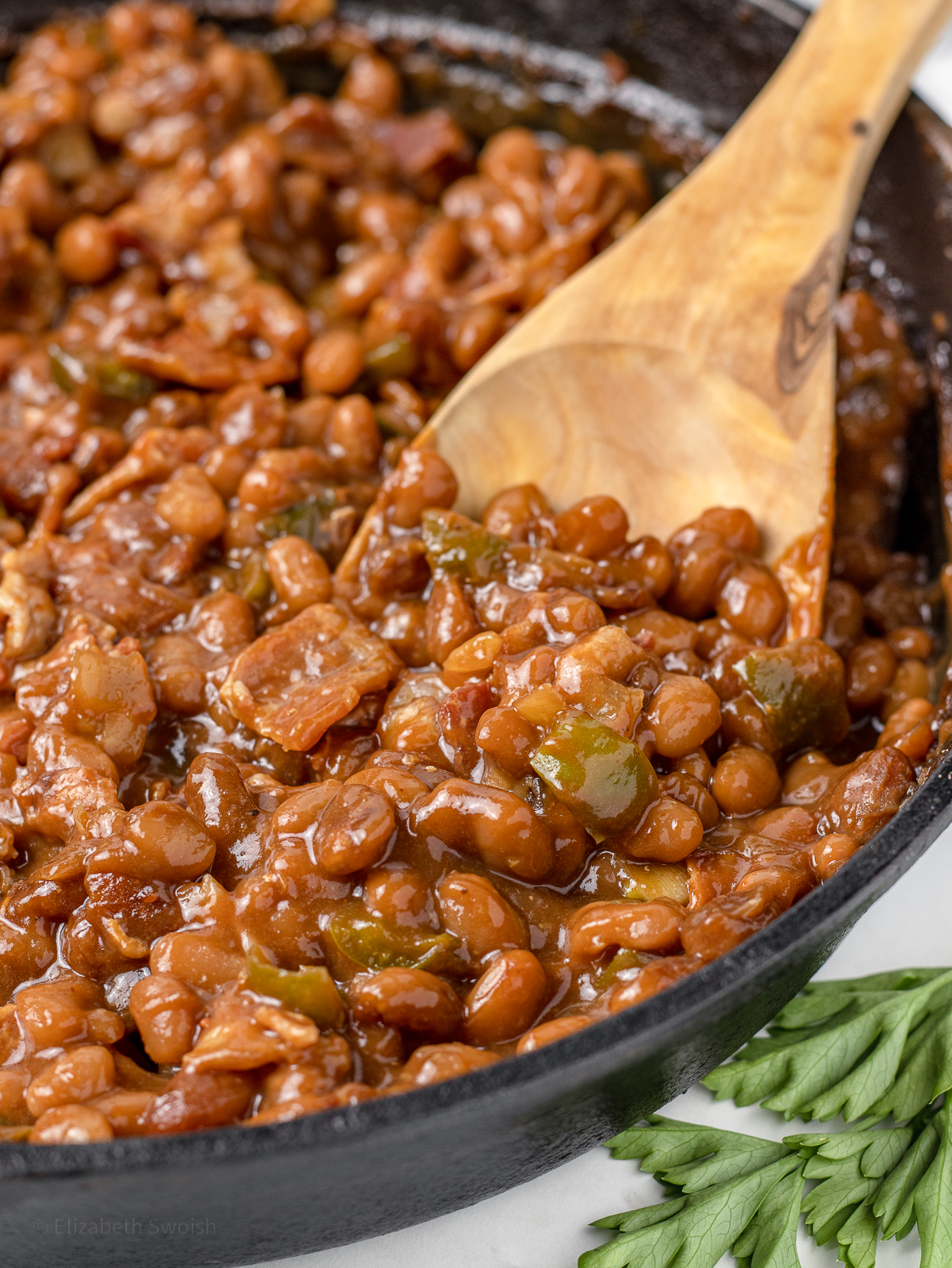Close up of Smoked Baked Beans in a cast iron skillet with bacon, onion, and green bell pepper with large serving spoon.