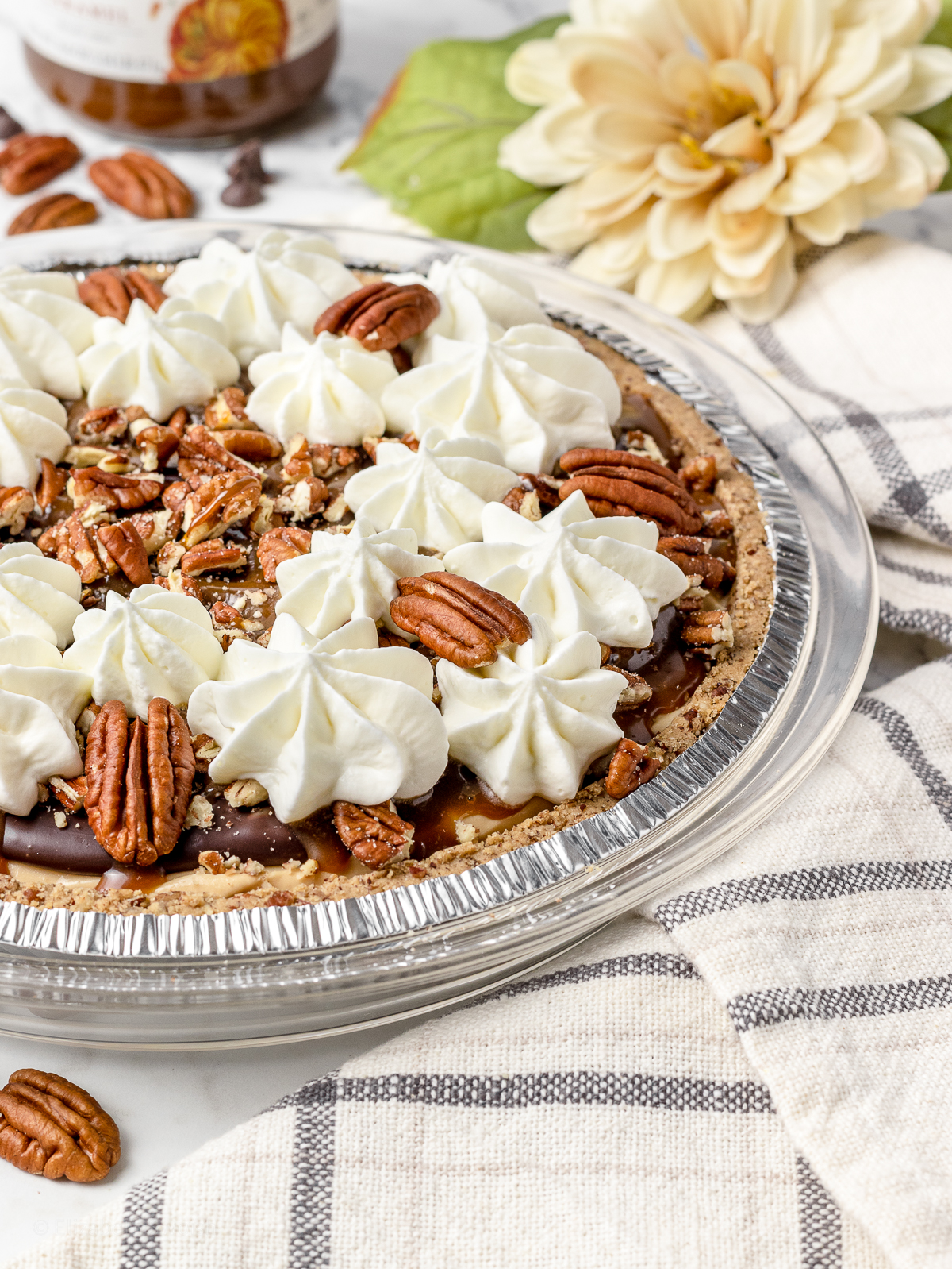 Close up of whole turtle pie to see toppings of chocolate ganache, caramel drizzle, whipped cream, chopped pecans, and pecan halves.
