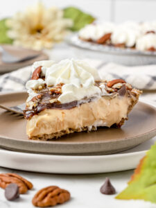 Slice of pie to show layers of pecan crust, caramel cream, chocolate ganache, caramel drizzle, chopped pecans, and whipped cream.
