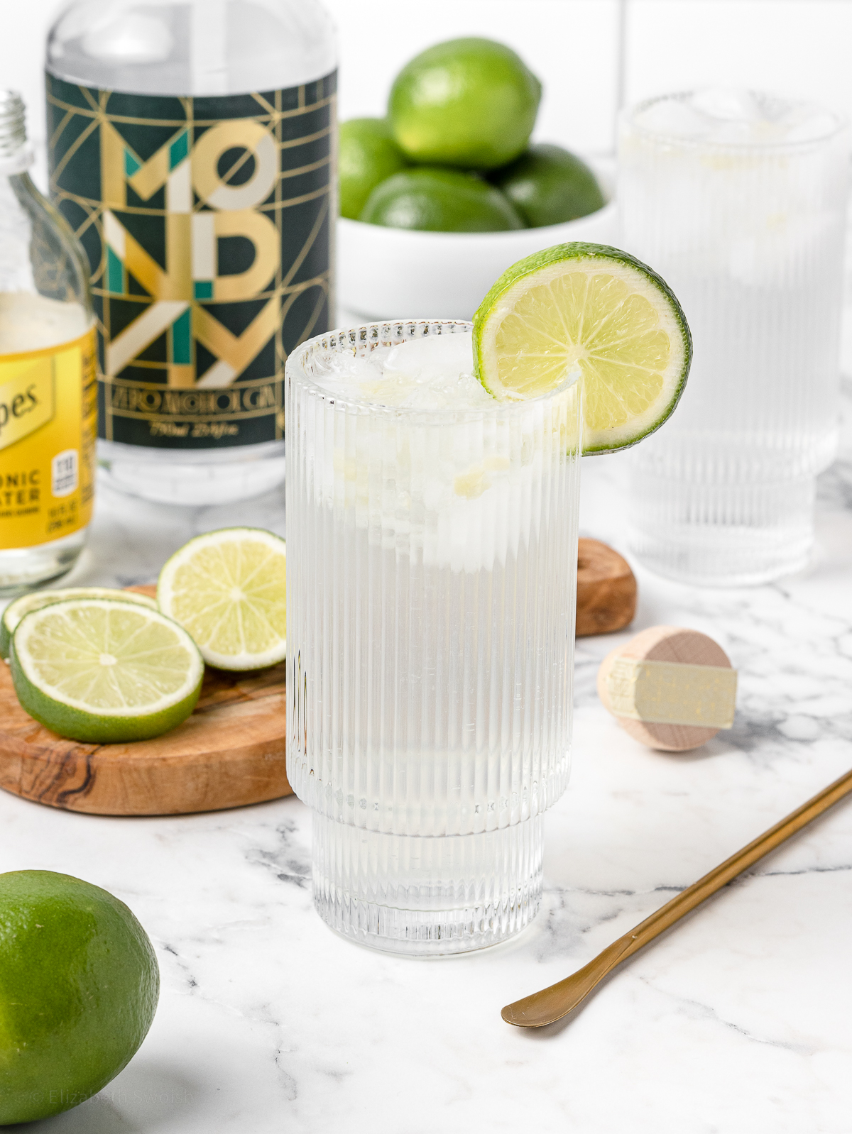 Gin and Tonic Mocktail in a highball glass with lime slice garnish. More limes and lime slices on the side. Non alcoholic gin, tonic water, and a stirrer in the background.