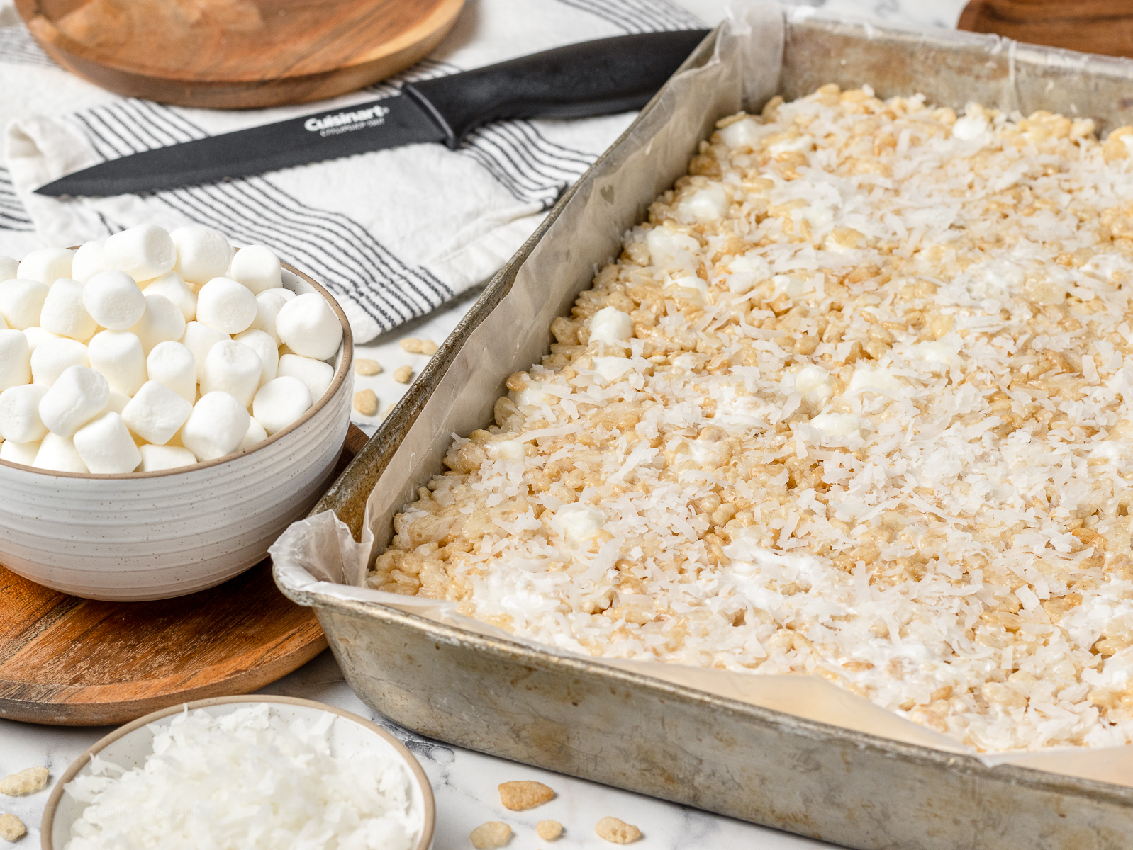 Coconut Rice Krispie Treats in a pan with the knife on the side ready to cut into squares. More sweetened coconut flakes on the side and mini marshmallows.