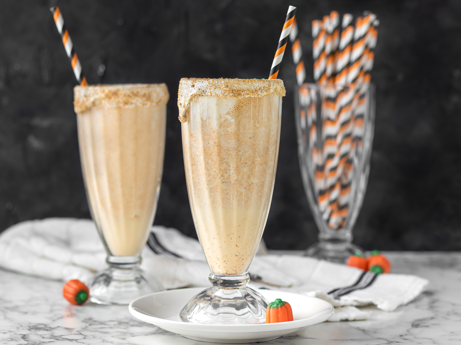 Two tall glasses of marshmallow and pumpkin flavored milkshakes with a orange and black striped straw and mini pumpkin candies on the side.