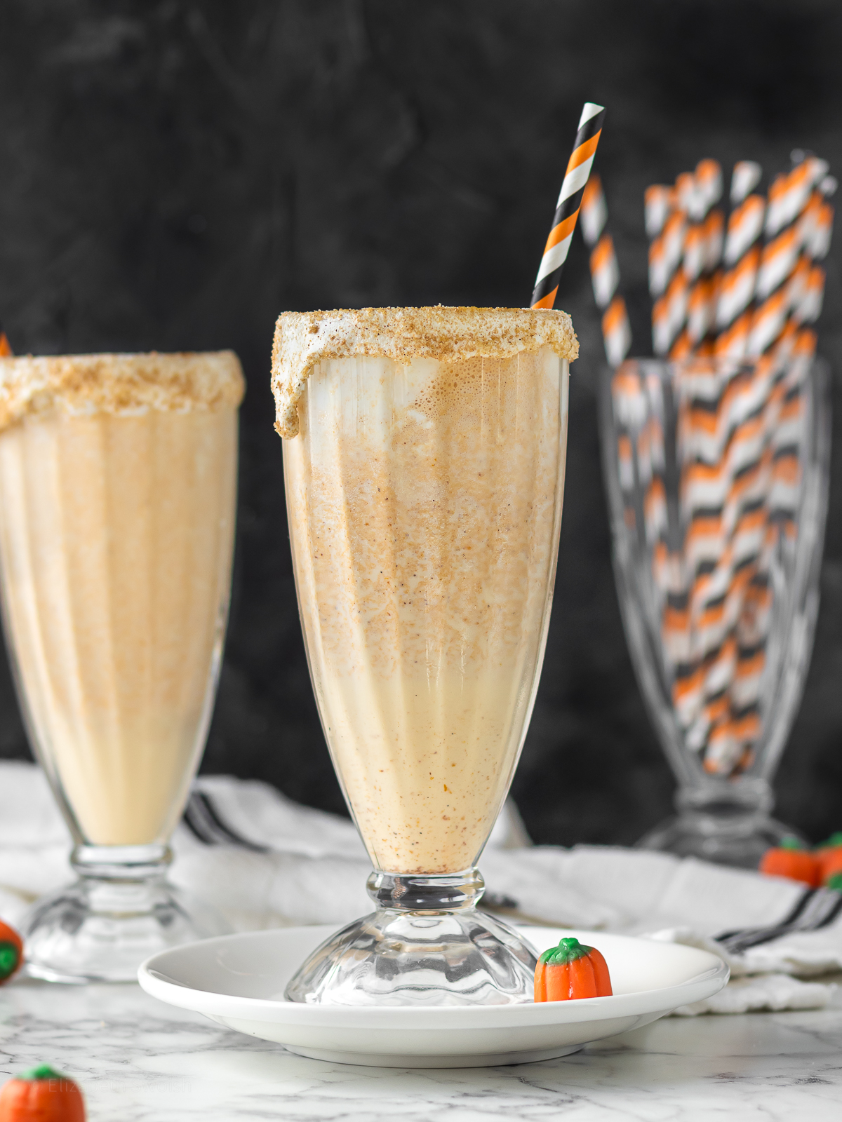 Two tall glasses of Marshmallow Pumpkin Milkshake with a orange and black striped straw and mini pumpkin candies on the side.