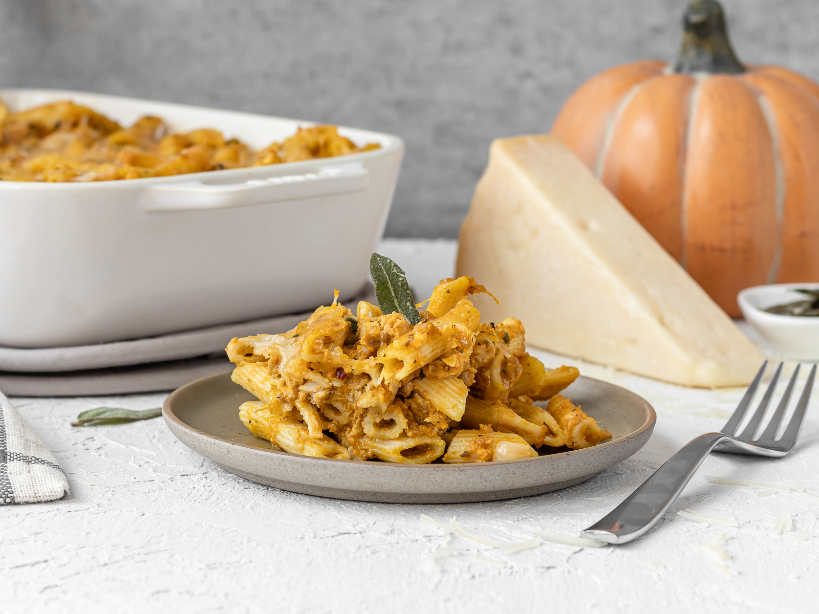 Plate of creamy pumpkin pasta topped with a fried sage leaf and parmesan cheese on the side with a baking dish filled with more pasta in the background.