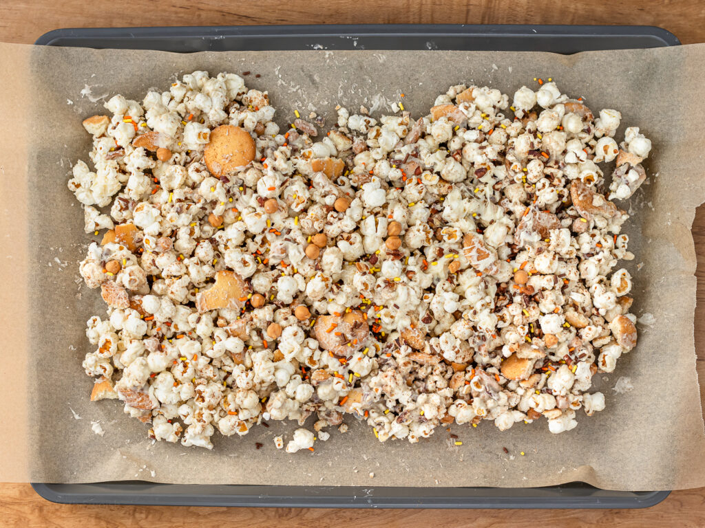 Sheet pan covered in parchment paper and the white chocolate pumpkin spice coated popcorn and goodies. Topped with cinnamon and festive fall sprinkles.
