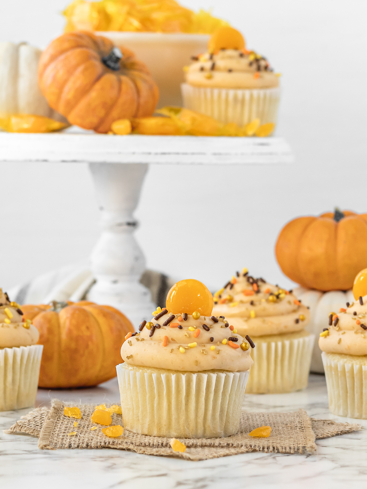 An arrangement of Butterscotch Cupcakes surrounded by orange pumpkins, white pumpkins, and butterscotch candies.