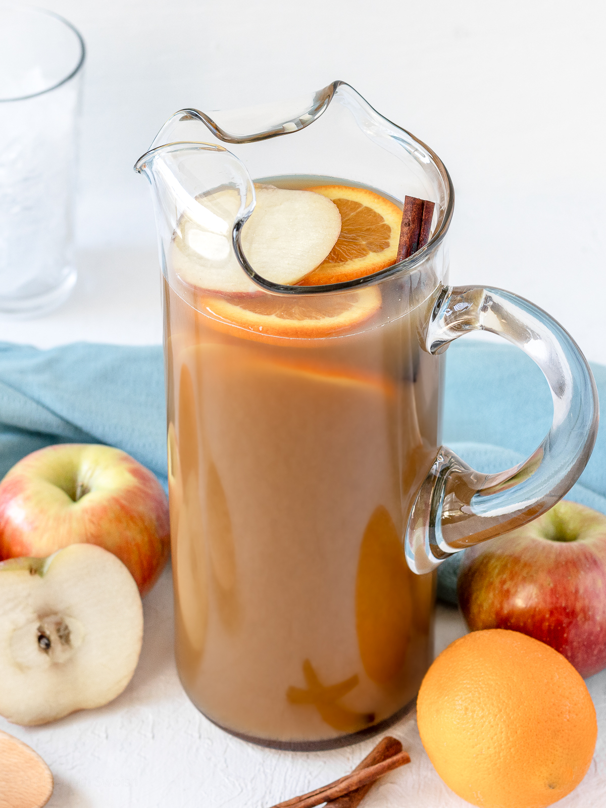 Large pitcher of non alcoholic Sweet Tea Harvest Punch surrounded by apples, oranges, cinnamon sticks, and glasses filled with ice.