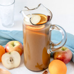 Large pitcher of non alcoholic punch surrounded by apples, oranges, cinnamon sticks, and glasses filled with ice.