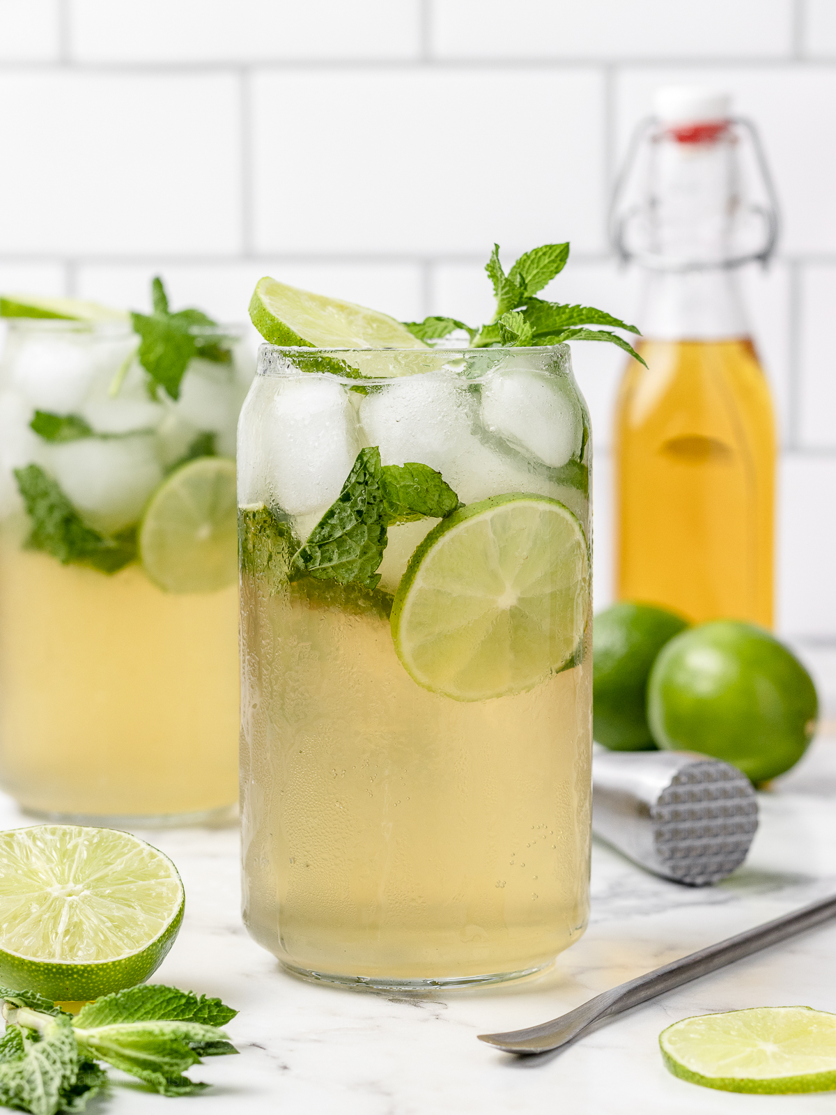 Two ice filled non alcoholic Mint Mojitos surrounded by limes, simple syrup, a muddler, and stirrer. Garnished with lime slices and fresh mint leaves.