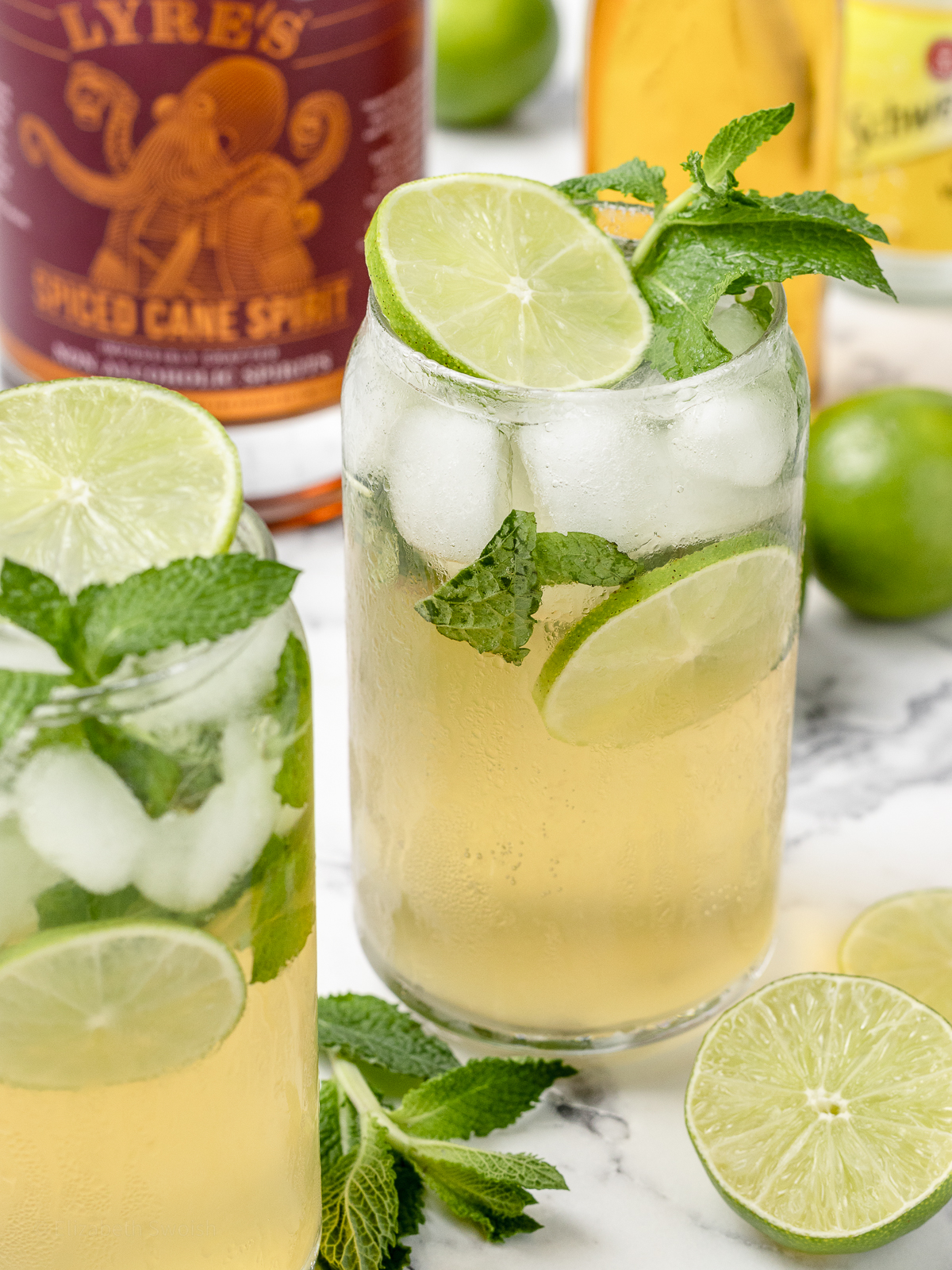 A Mint Mojito Mocktail garnished with lime slices and mint leaves. A bottle of zero proof spiced rum, agave syrup, and tonic water on the side.