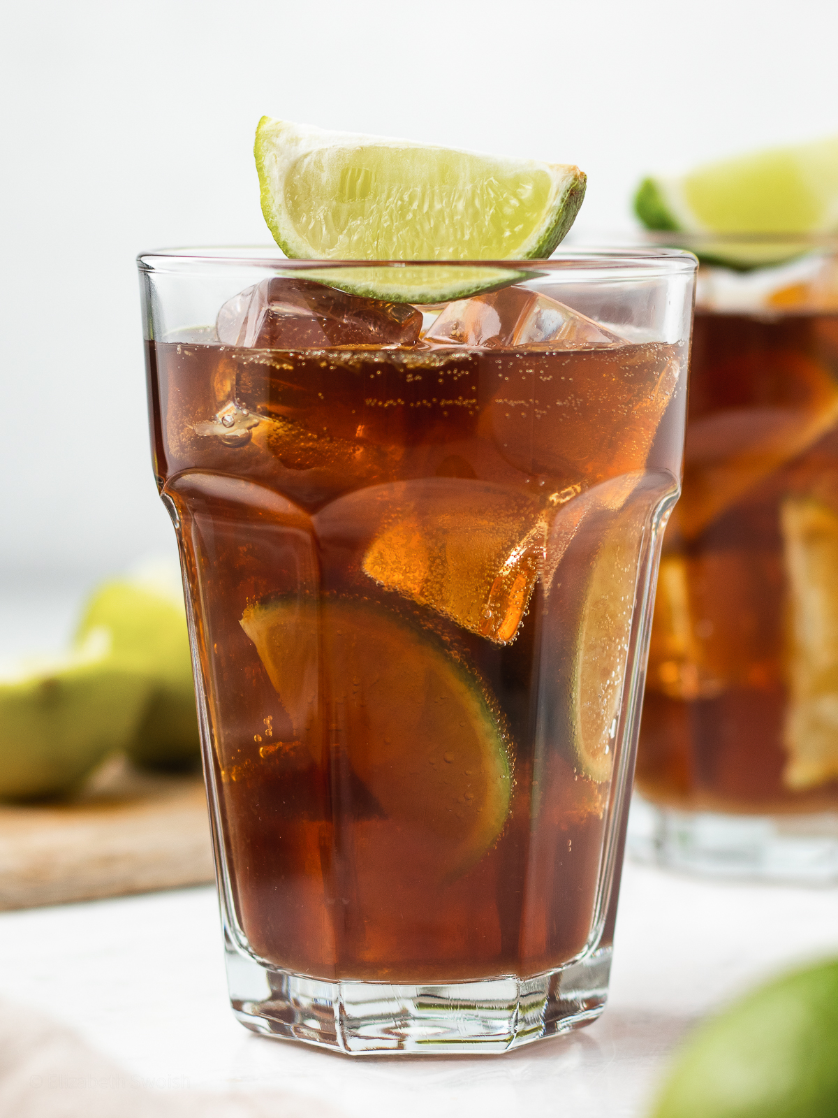 Two non alcoholic Cuba Libre drinks with a lime wedge garnish on top and more on the side.