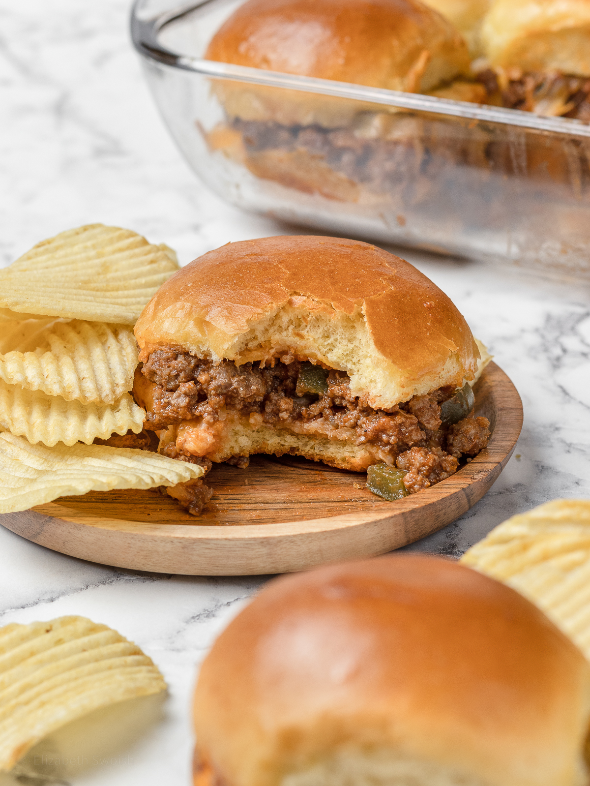 Sloppy joe that has one bite taken from it and chips on the side.