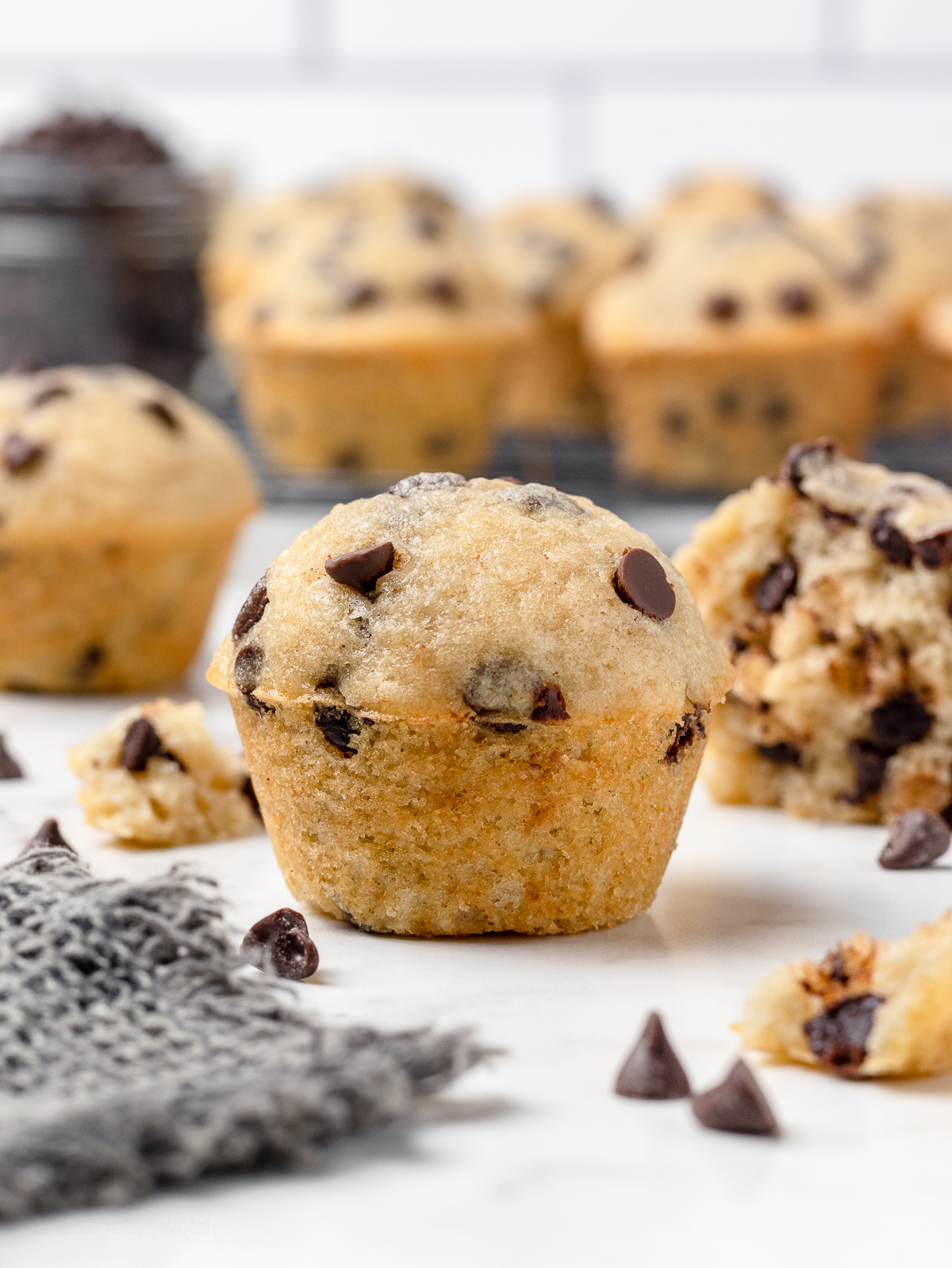 A tall mini muffin with chocolate chips and more mini muffins on a cooling rack in the background.