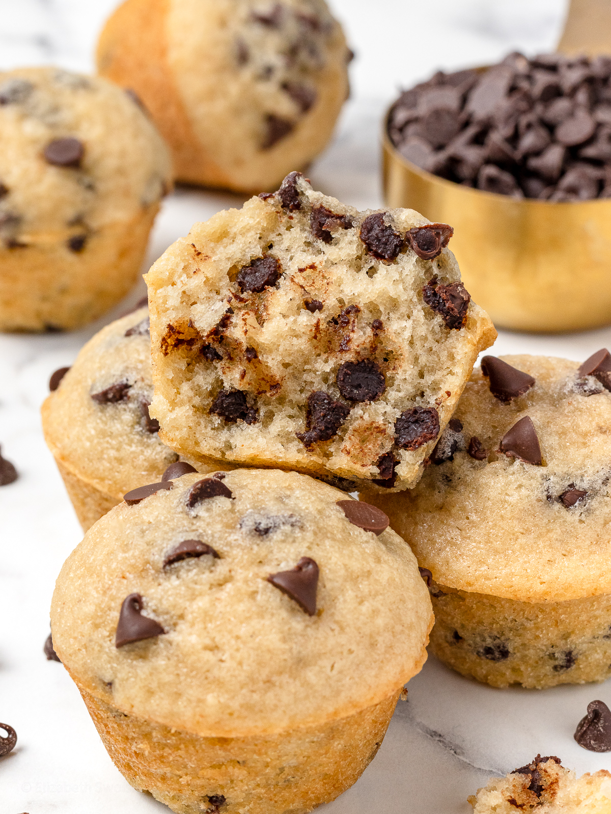 A pile of mini chocolate chip muffins with a scoop of mini chocolate chips in the background.