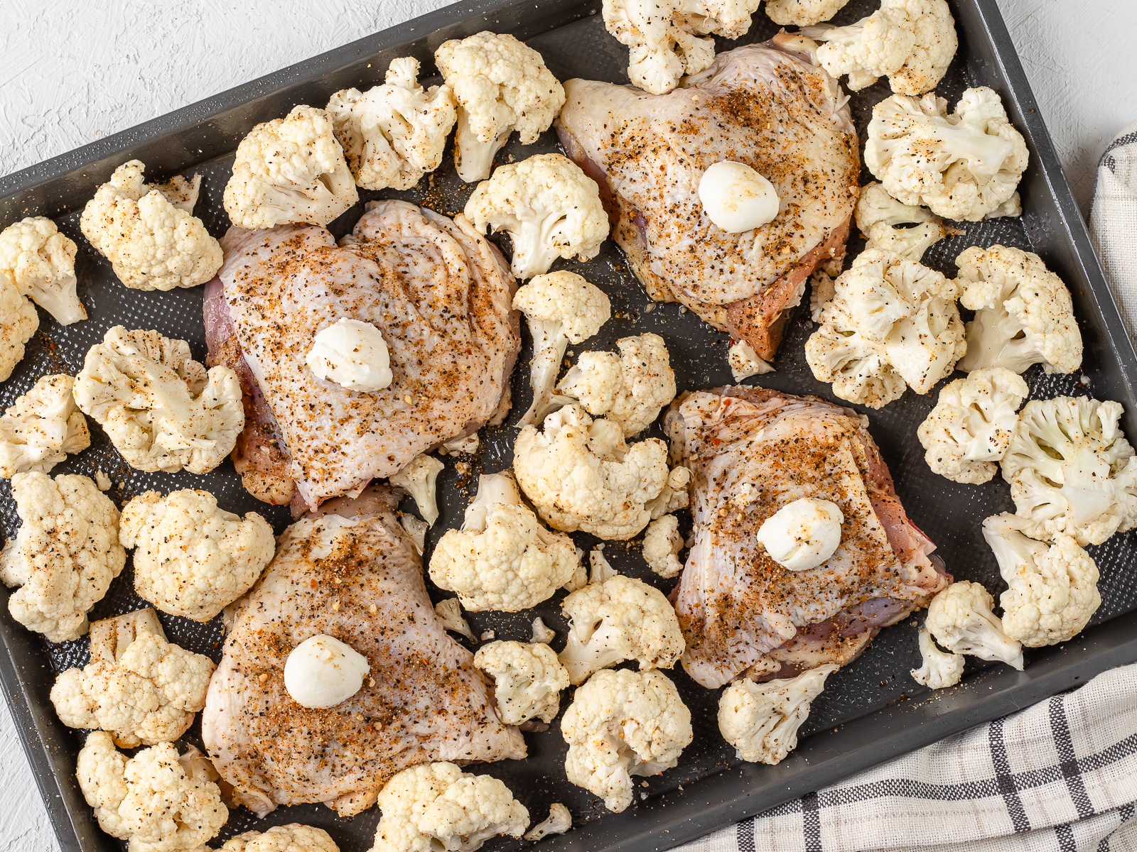 A baking sheet topped with a dollop of butter and cajun seasoned chicken thighs. Seasoned cauliflower surrounding the chicken and ready to go in the oven.