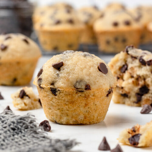 A tall mini muffin with chocolate chips and more mini muffins on a cooling rack in the background.