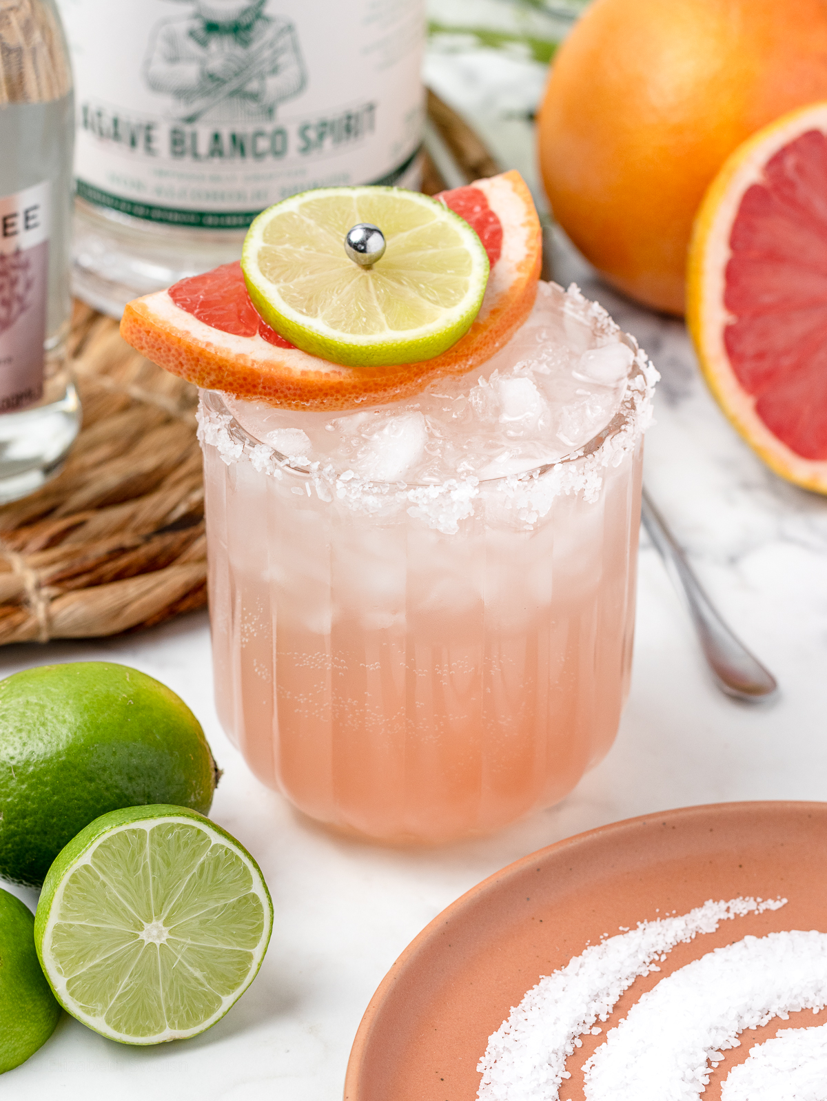 A Paloma Mocktail with ice, salted rim, and garnished with lime and grapefruit. The drink is surrounded by it's ingredients and a long handled stirrer.