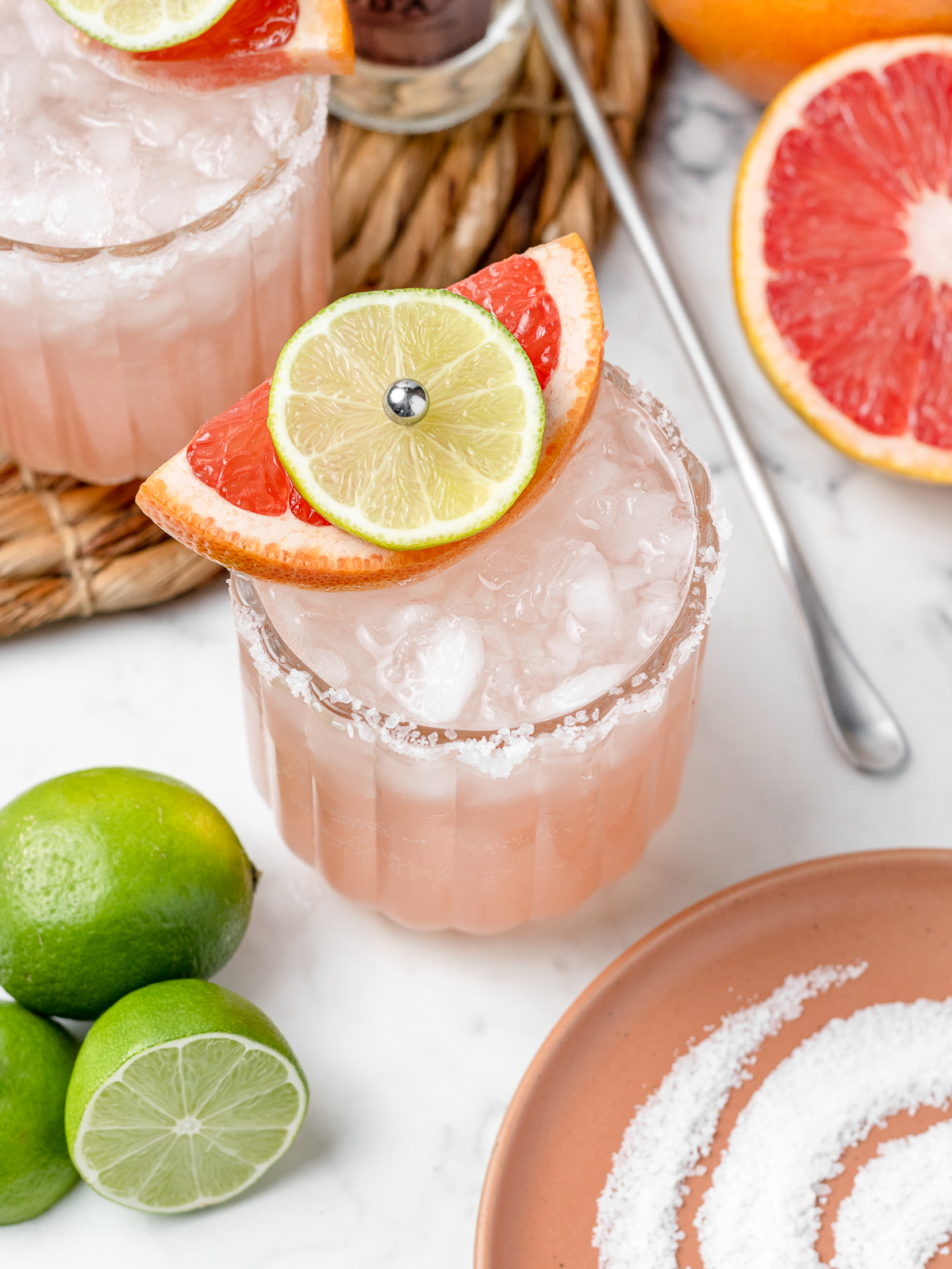 Two Paloma Mocktails with grapefruit and limes around them. There is also a plate of salt for the rim of the glass and a long handled stirrer.