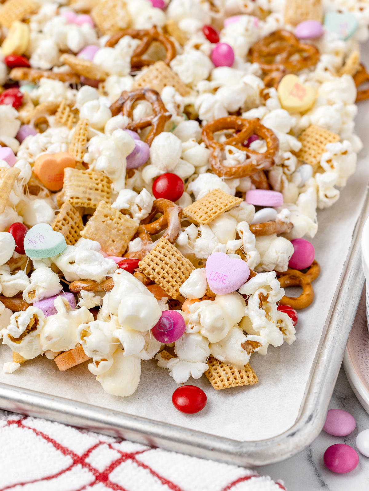 Valentines Day themed popcorn snack mix with mini pretzels and corn chex, coated with marshmallow, and topped with M&M's and conversation hearts.