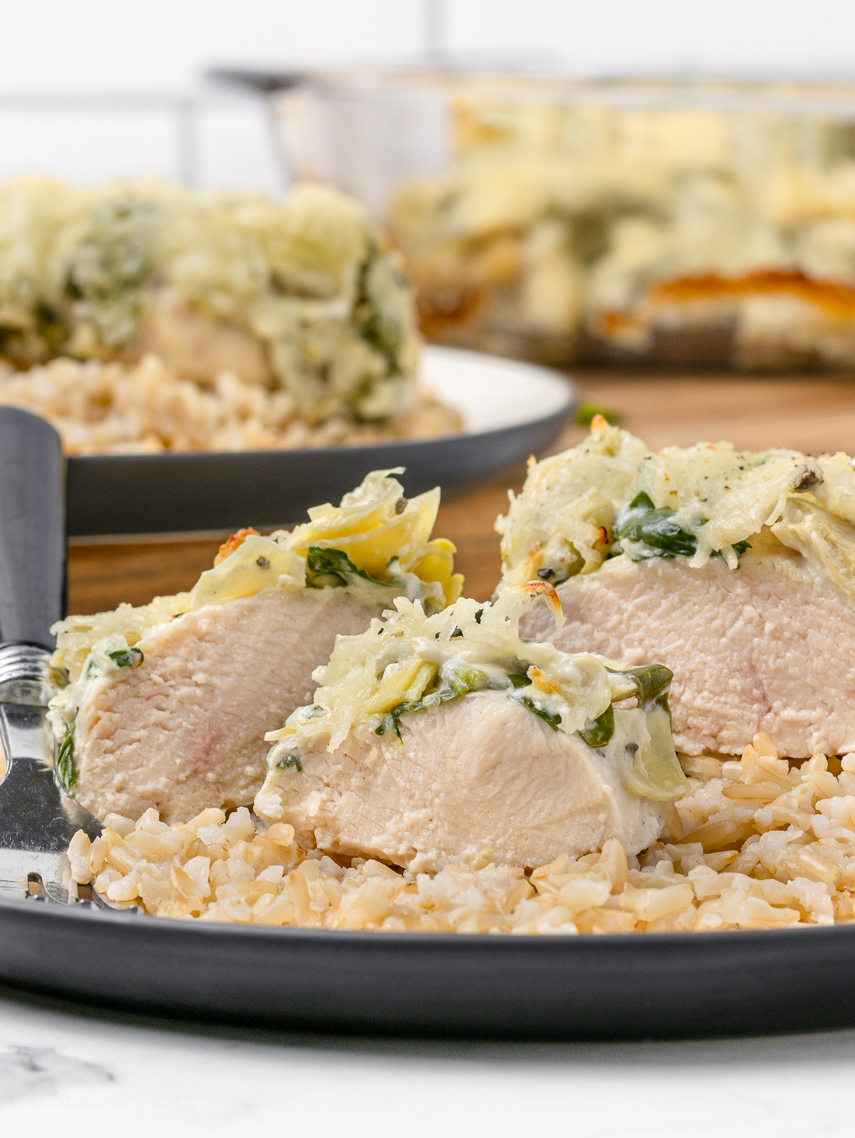 A plate with cut chicken on a bed of brown rice. The chicken is moist and topped with a creamy and cheesy spinach artichoke topping. Large casserole dish of more chicken in the background.