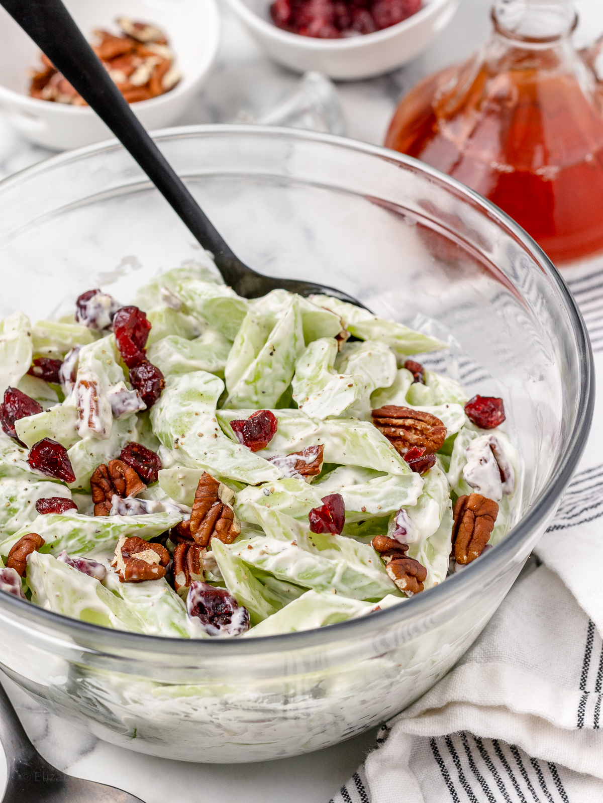 A big bowl of Celery Salad and a spoon ready for serving. More chopped pecans, dried cranberries, and a bottle of red wine vinegar in the background.