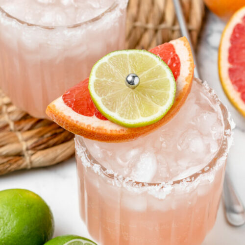 Close up of a non alcoholic paloma and it's garnish. Another drink is in the background with more limes and grapefruits on the side.