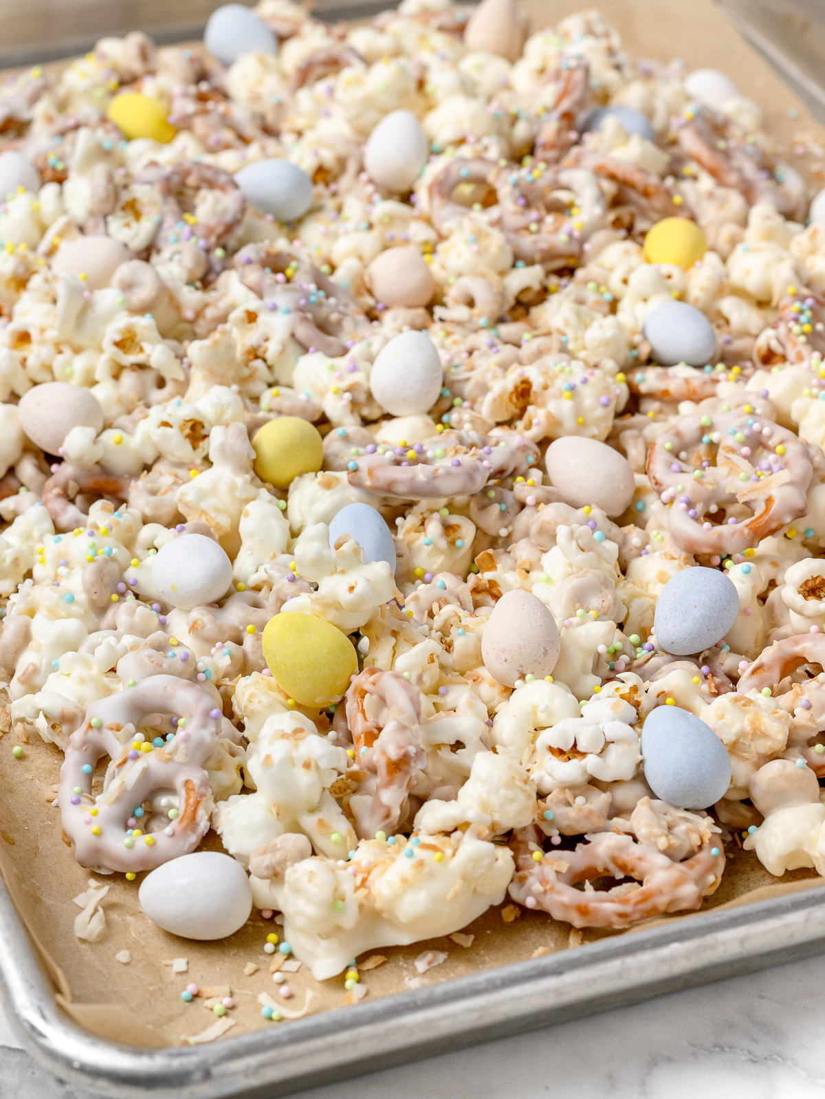 Popcorn Snack Mix on a baking sheet topped with toasted coconut, milk chocolate candy eggs, and pastel Easter sprinkles.