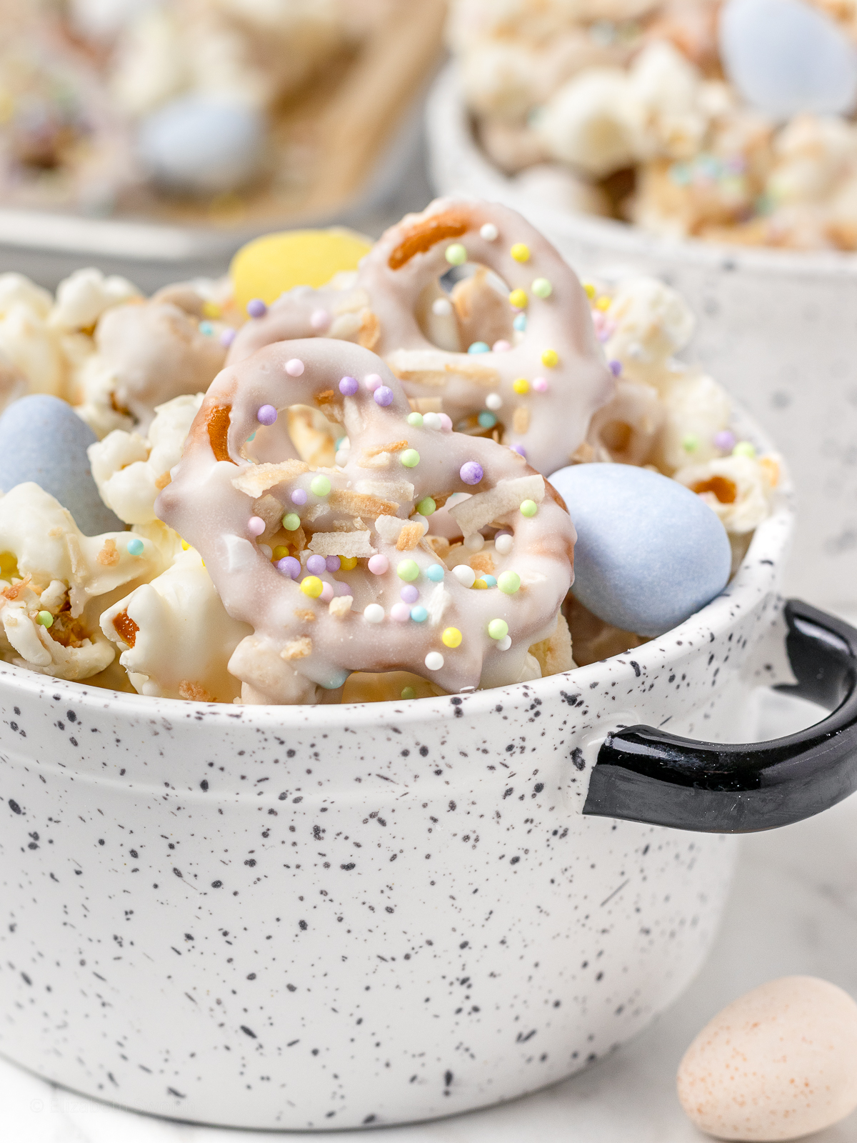 A bowl of Bunny Popcorn Snack Mix. Close up of a white chocolate covered pretzel with toasted coconut and pastel Easter sprinkles stuck to it. Colorful milk chocolate candy eggs and more bowls of popcorn snack mix on the side.