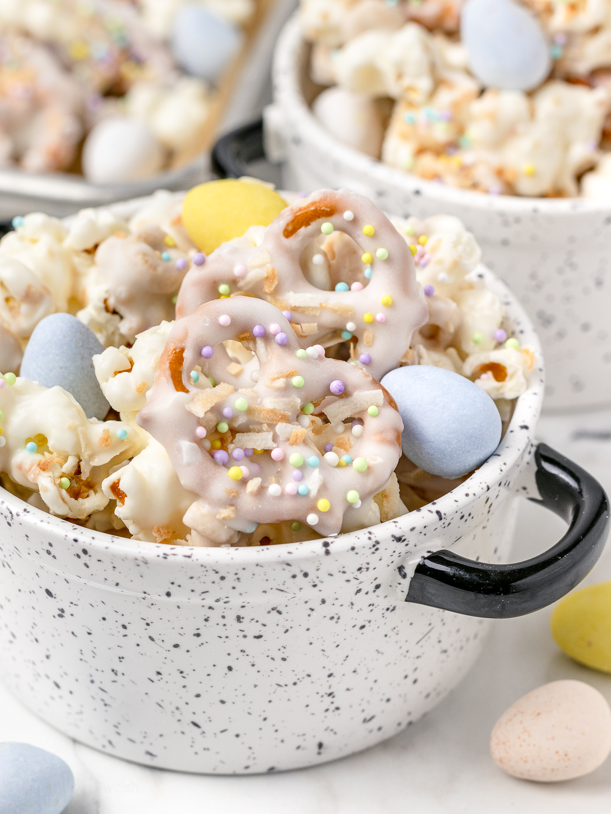 A bowl of Bunny Popcorn Snack Mix with white chocolate coated popcorn, pretzels, toasted O's cereal topped with milk chocolate candy eggs, and pastel Easter sprinkles.