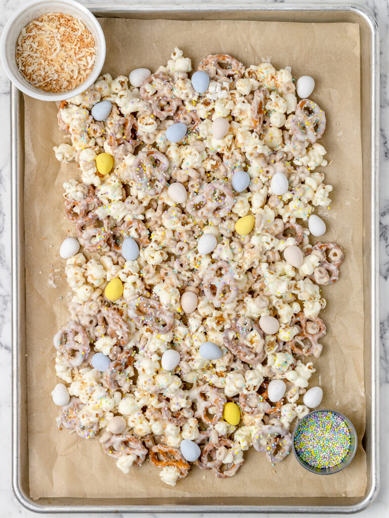 Chocolate coated popcorn snack mix topped with unsweetened toasted coconut, milk chocolate eggs, and pastel Easter sprinkles on a parchment paper lined baking sheet. Waiting for the chocolate to harden.