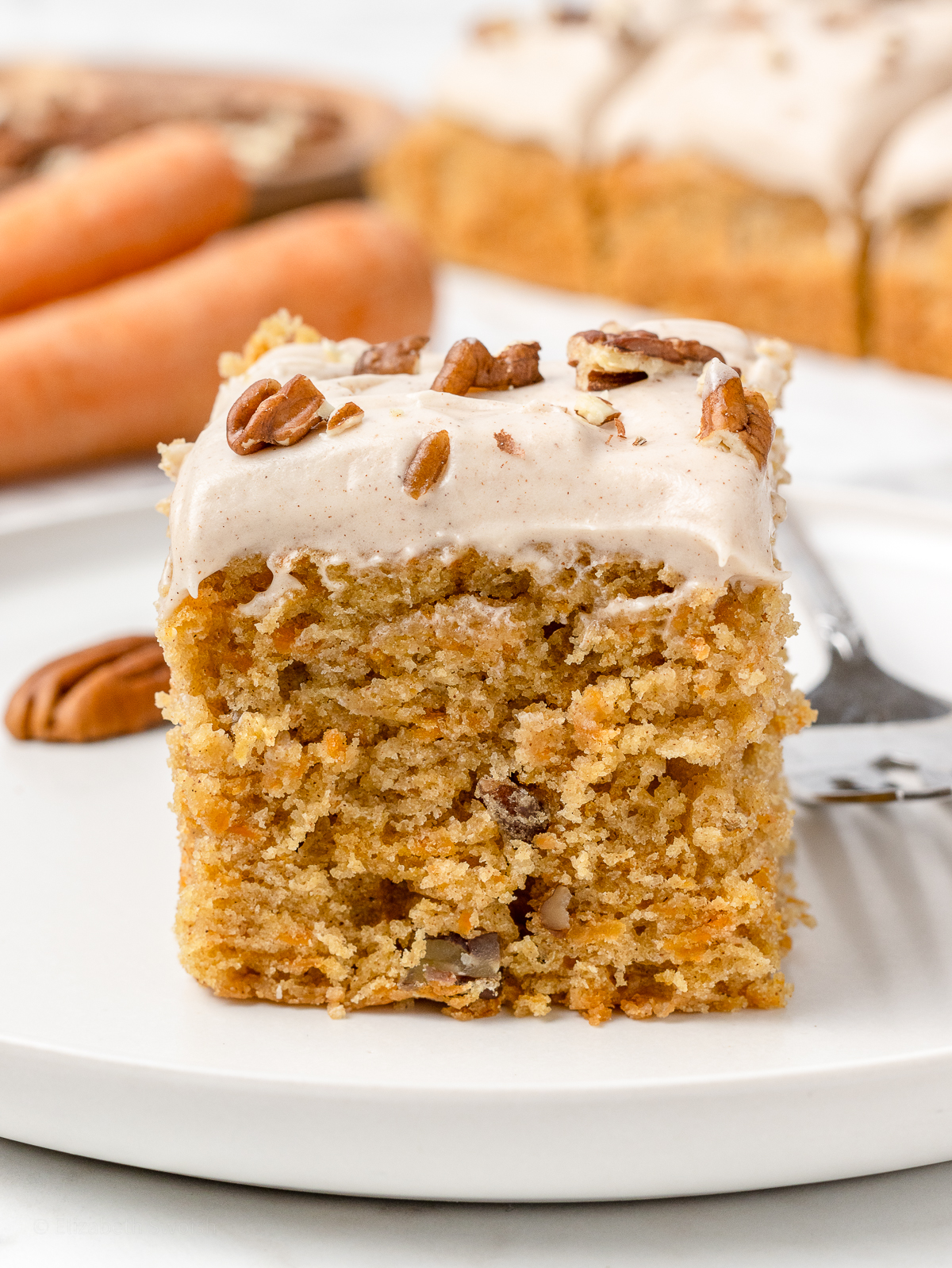 A slice of carrot snack cake on a plate with a fork on the side. It is ready to eat.