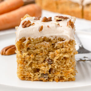 A slice of carrot snack cake topped with cinnamon cream cheese buttercream and toasted pecans on a plate with a fork on the side.