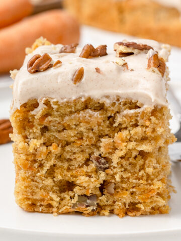 A slice of carrot snack cake topped with cinnamon cream cheese buttercream and toasted pecans on a plate with a fork on the side.