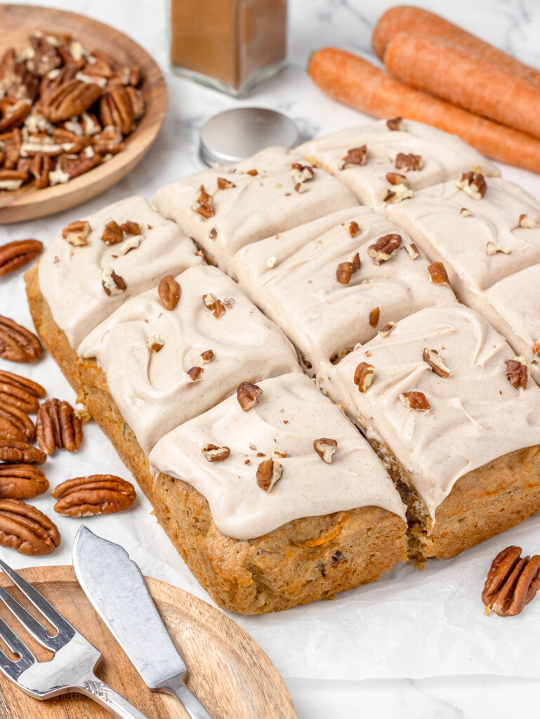 A one layer carrot cake cut into 9 slices ready to serve. More toasted pecans and ground cinnamon on the side.