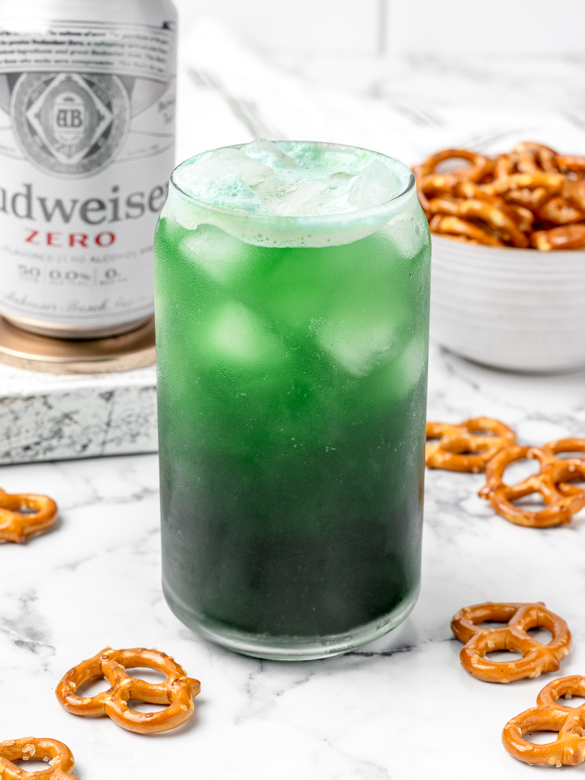 A non alcoholic green beer with another beer and pretzels in the background.