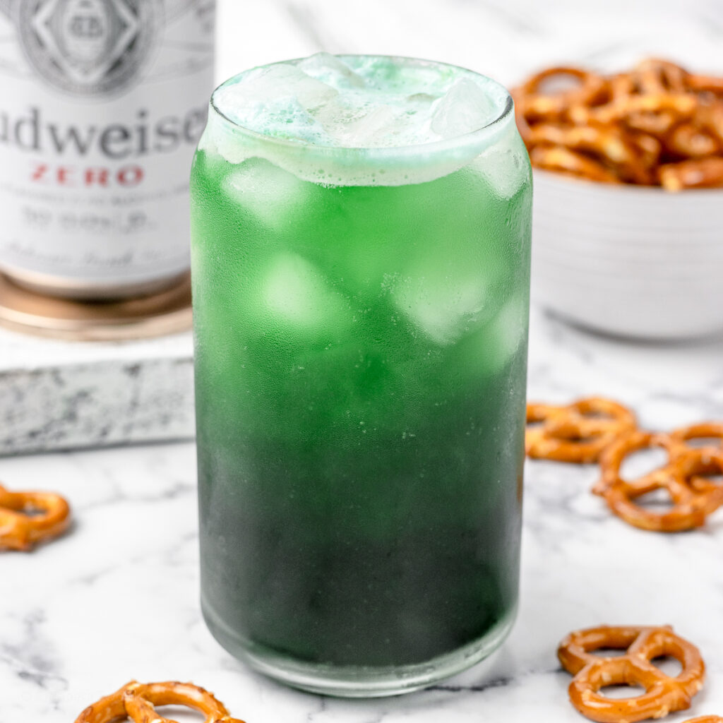 A green beer with non alcoholic beer and pretzels in the background.