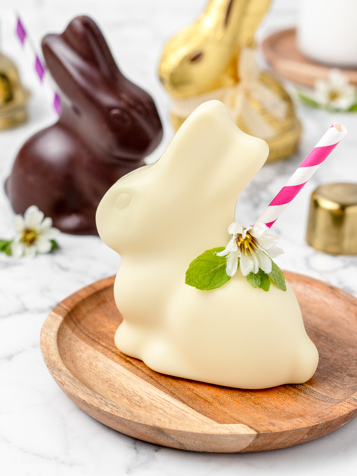 2 Chocolate Bunny Easter Mocktails garnished with a straw, mint leaves, and mini edible flour. One mocktail is white chocolate, another is milk chocolate, and there is a gold wrapped chocolate bunny in the background.