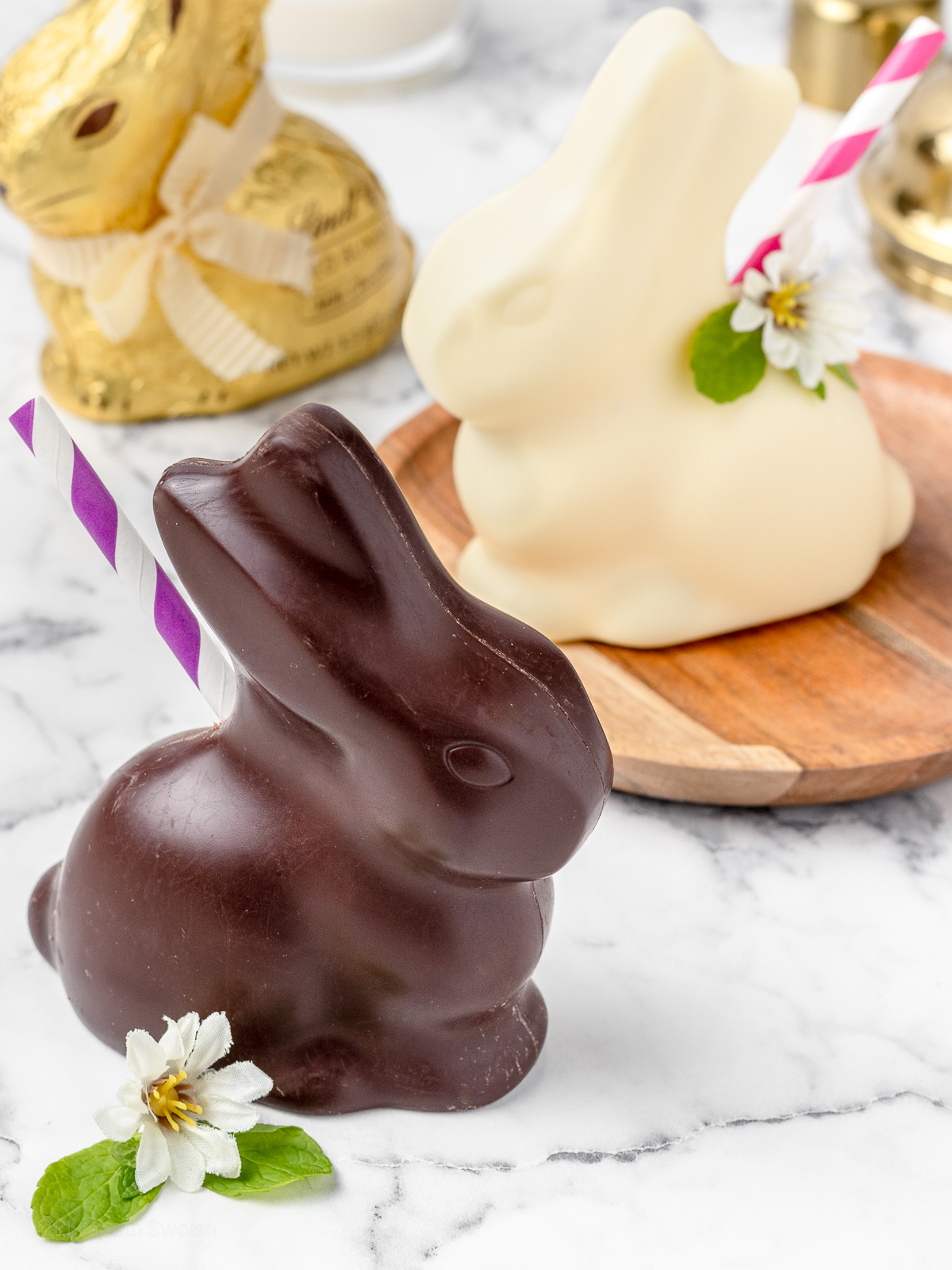 A milk chocolate bunny mocktail with a white chocolate bunny mocktail in the background. They are garnished with colorful striped straws, mint leaves, and edible flowers.