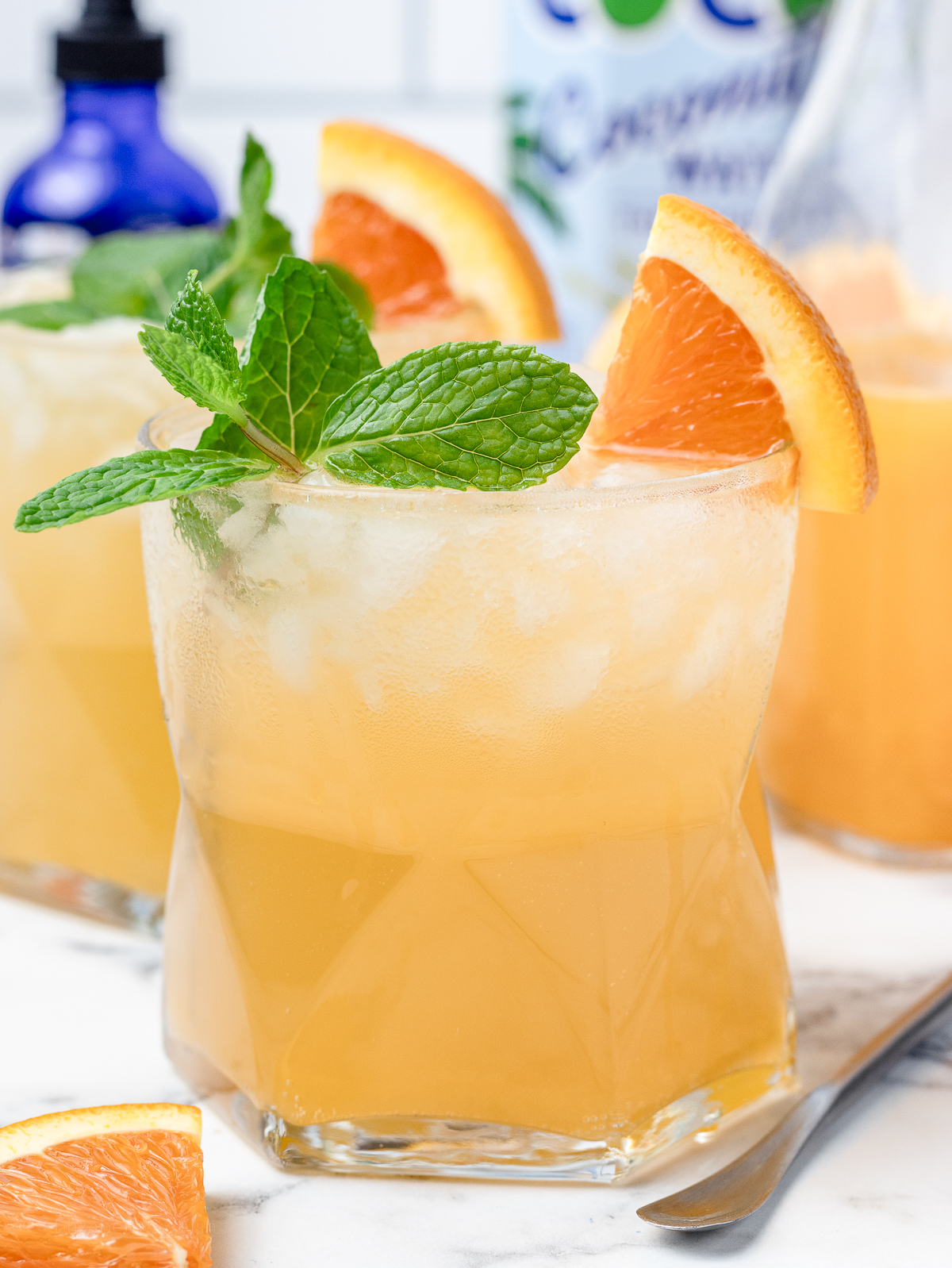 An iced Orange Coconut Water Mocktail with fresh mint and orange slices. They are a light and refreshing orange color. There is freshly squeezed orange juice, alcohol free orange bitters, and coconut water in the background.
