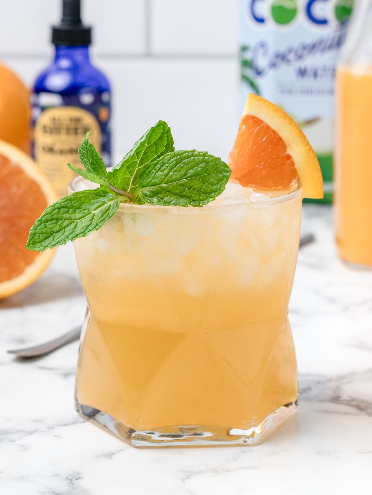 A refreshing coconut and orange mocktail with orange bitters, coconut water, and freshly squeezed orange juice in the background.
