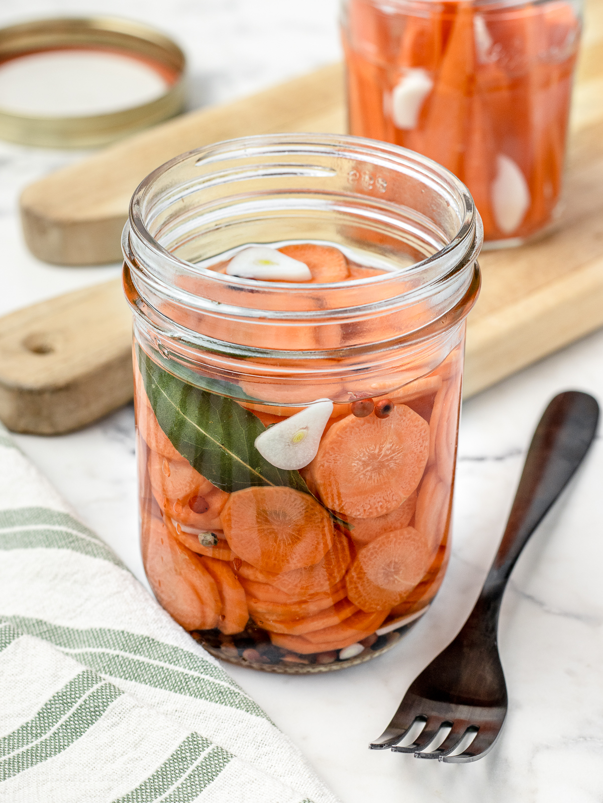Two jars of quick pickled carrots seasoned with garlic cloves, peppercorns, and bay leaves. One jar has carrot sticks, the other has sliced carrots.