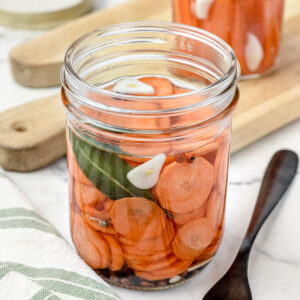 Jar of quick pickled carrots seasoned with garlic cloves, peppercorns, and bay leaves. One jar has sliced carrots and the other has carrot sticks.