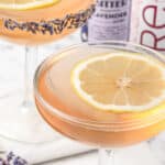 Close up of Lavender Mocktail with a lemon slice floating on top and a lavender bud rim. Lavender Bitters and sparkling rose in the background.