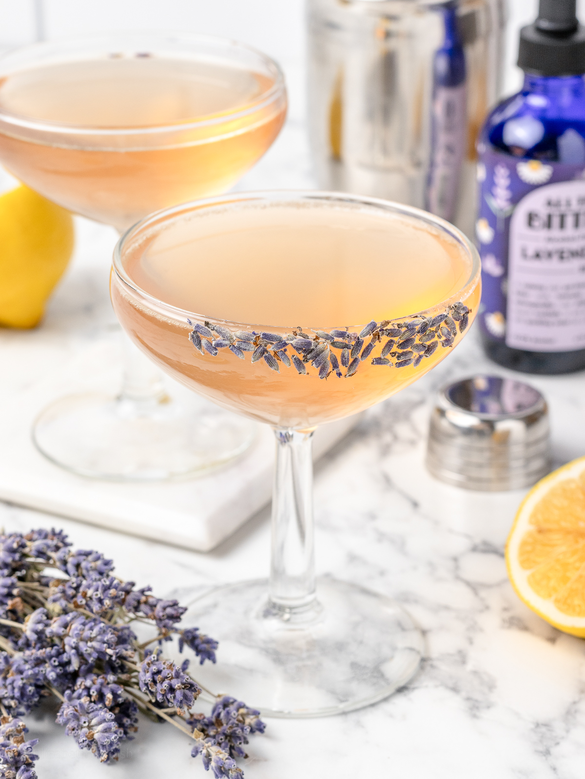 Two coupe glasses filled with the Sparkling Lavender Mocktail. One glass is garnished with a lavender bud rim and more lavender flowers on the side.