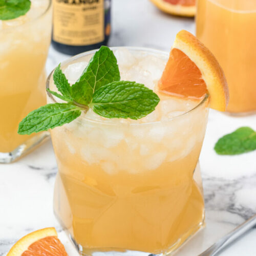 A fresh Orange Coconut Water Mocktail with mint and orange slices. A stirrer, more orange juice, and another mocktail on the side ready for sipping.